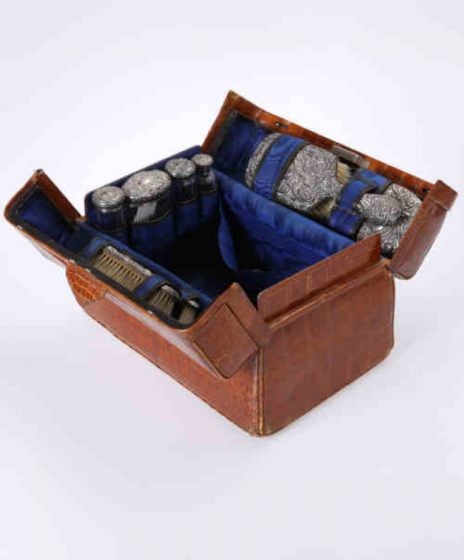 An Edwardian Alligator skin dressing case, with blue silk interior, fitted with silver and glass bottles, silver backed brushes and ivory glove stretchers, H. Matthews, the majority Birmingham 1901.