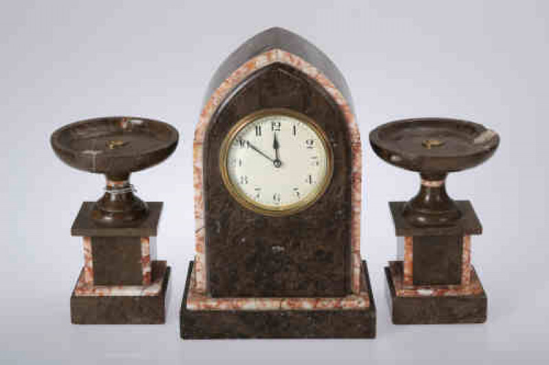 An Art Deco two tone marble clock garniture, the clock with pointed arch case and plinth base, the white enamel dial with Arabic numerals, within a brass bezel with bevelled glass, the cylinder movement with slow/fast switch, the pair of urns with integra