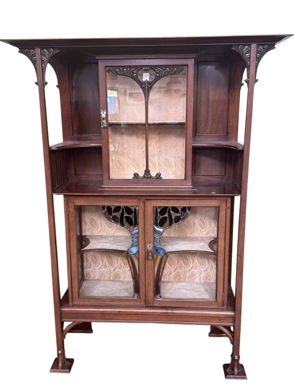 Art Nouveau mahogany cabinet, the canopied top having central glazed panel door flanked by two shaped shelves above two coloured leaded glass panel doors, 183.5cm by 138cm by 46cm.