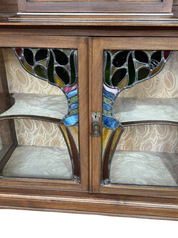 Art Nouveau mahogany cabinet, the canopied top having central glazed panel door flanked by two shaped shelves above two coloured leaded glass panel doors, 183.5cm by 138cm by 46cm.