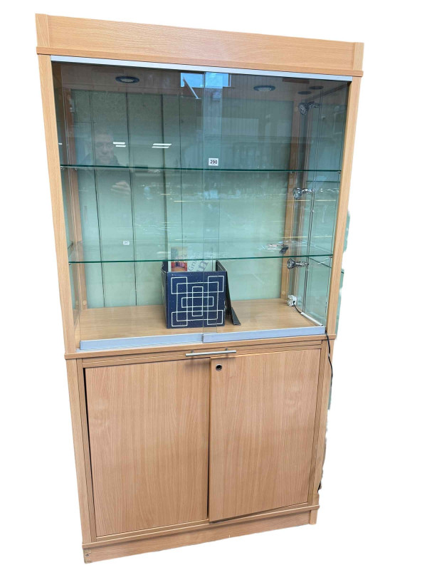 Beech finish illuminated shop display cabinet having two sliding glazed doors above a slide with two cupboard doors below, 191cm by 99.5cm by 49.5cm.