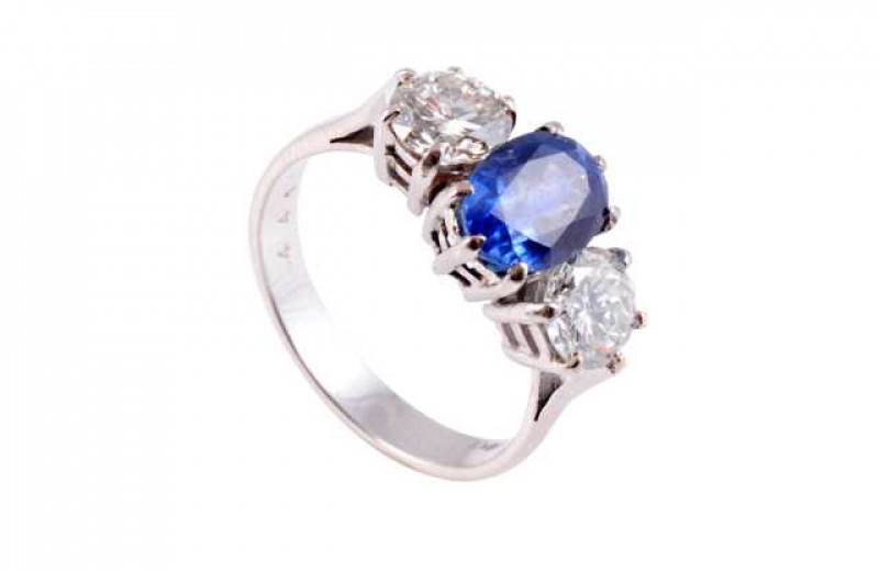 A sapphire and diamond ring, the central oval cut sapphire six claw set between two brilliant cut five claw set diamonds, all set in a pierced mount between open tapering shoulders, on a plain band. Stamped 18KT. Ring size N1/2