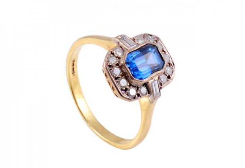A sapphire and diamond ring, set to centre with a rectangular cut sapphire collet set with a bezel of twelve old brilliant cut claw set diamonds and two baguette cut diamonds, collet set, horizontal to each shoulder, all set in pierced detail mount betwee