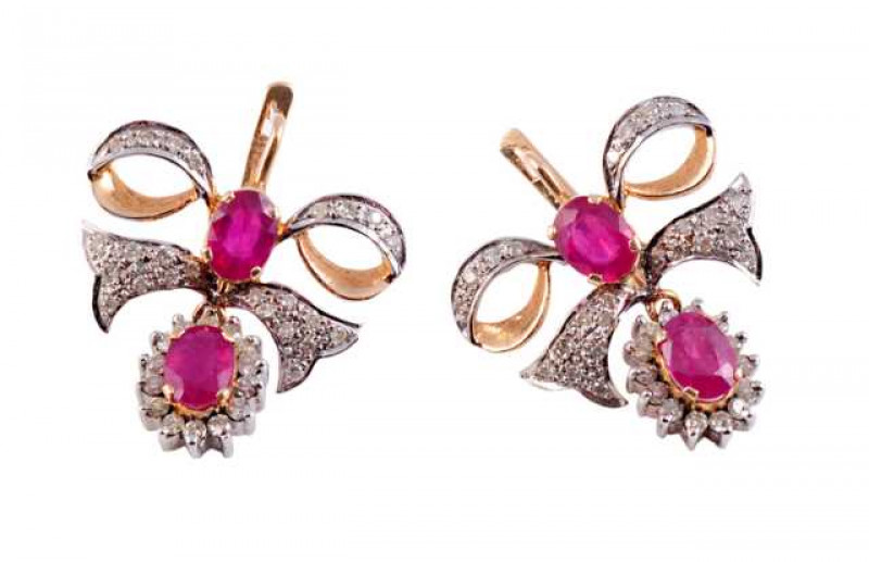 A pair of ruby and diamond earrings, the oval cut ruby four claw set within a surround of fourteen brilliant cut claw set diamonds, all suspending on an articulated link from the bottom of a stylised bow design, set to the centre with a similar oval cut f