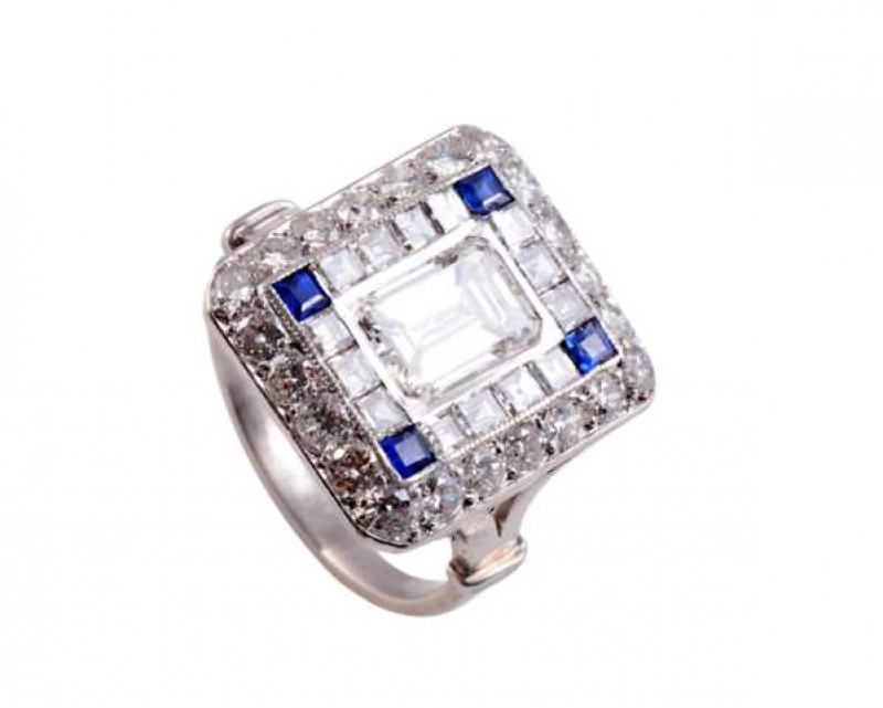 A diamond and sapphire ring, the square shaped mount set to the centre with an emerald cut diamond, collet set within an inner bezel of fourteen princess cut channel set diamonds and four channel set square cut sapphires to each corner, in turn all set wi