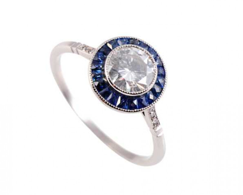 A sapphire and diamond ring, the central brilliant cut diamond millegrain set within a bezel of twenty one square cut sapphires, all set in a pierced detail open mount set between open shoulders each set with three brilliant cut diamond highlights, on a p