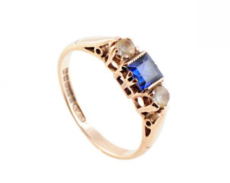 A sapphire and diamond ring, the central square cut claw set sapphire set between a brilliant cut claw set diamond to each side, all set in a pierced detail mount between similar pierced detail tapering shoulders on a plain band. Stamped 375. Makers mark 