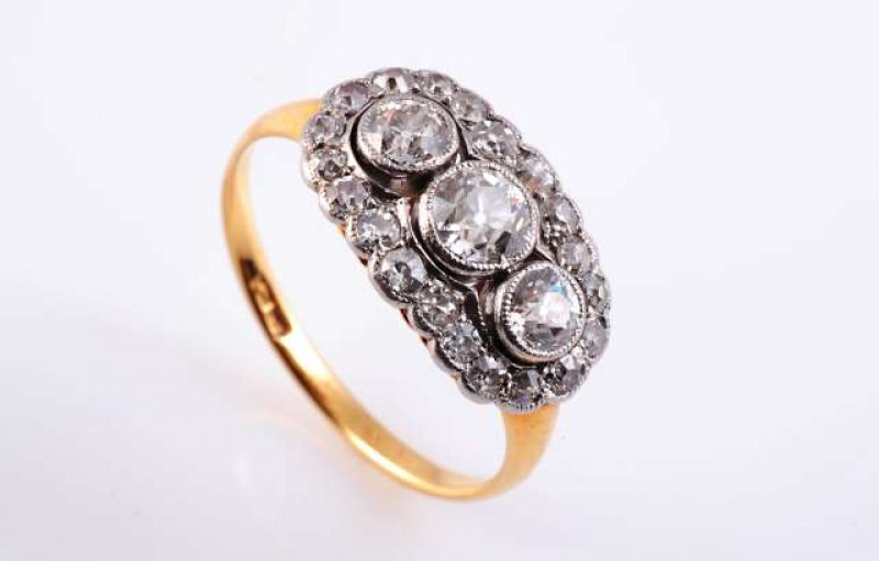A DIAMOND CLUSTER RING c.1900-1910 The three graduating old brilliant cut millegrain set diamonds set to the centre of a scalloped edge mount set with twenty slightly graduating round mixed cut millegrain set diamonds, all set in a pierced open mount betw