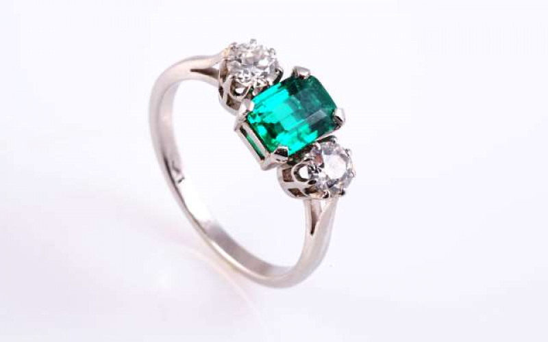 AN EMERALD AND DIAMOND RING The rectangular cut-cornered emerald four claw set between two brilliant cut eight claw set diamonds, all set in a pierced detail mount between open tapering shoulders on a plain band. Stamped PLAT (hallmark very worn). Ring si