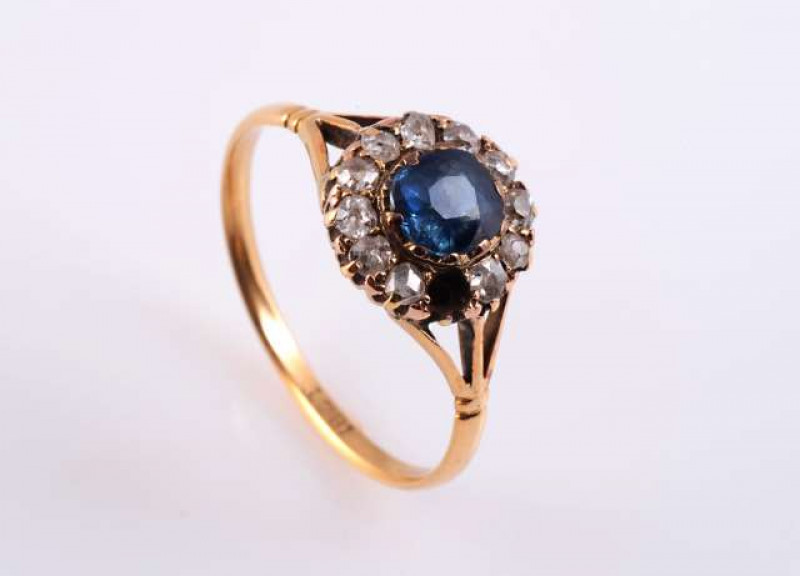 A SAPPHIRE AND DIAMOND RING c.1890-1900 The round mixed cut sapphire claw and collet set within a surround of twelve rose cut claw set diamonds (one diamond lacking), all set in an open mount between split design shoulders with moulded detailing on a plai