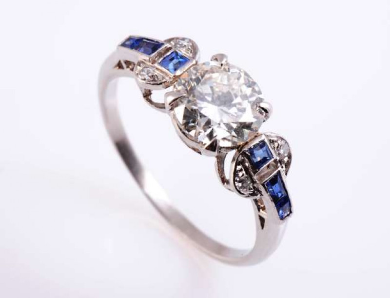 AN ART DECO SAPPHIRE AND DIAMOND RING The brilliant cut diamond four claw set between moulded circular shoulders each set with two brilliant cut diamond highlights and three square cut channel set sapphire highlights, all set in an open pierced mount betw