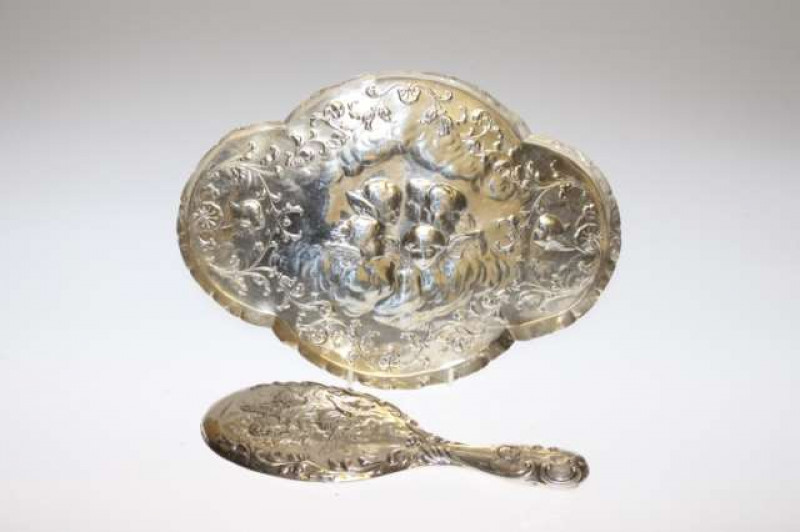 AN EDWARDIAN SILVER DRESSING TABLE TRAY, William Comyns, London 1905, of elongated quatrefoil form, embossed with winged cherub masks and scrolling foliage; together with A SILVER BACKED MIRROR, London 1901, similarly decorated but by a different maker. (