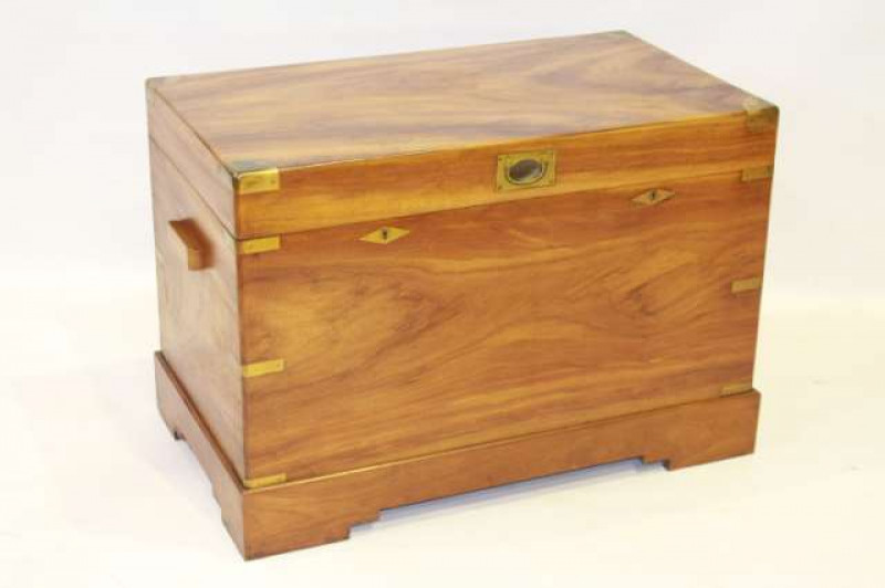 A BRASS-BOUND TEAK CAMPHOR CHEST, with brass 'campaign' handles and twin locks with brass escutcheons, raised on a plinth stand. 65cm by 95cm by 55cm