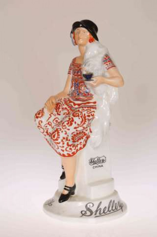 SHELLEY GIRL', A SHELLEY POTTERY ADVERTISING FIGURE, modelled seated on a pedestal holding a tea cup and wearing a blue and coral red paisley dress, printed Shelley to either side of the pedestal and twice to the base, green printed factory mark to unders