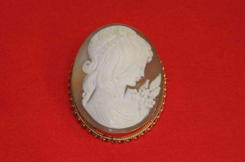 A cameo brooch, the oval cut carved cameo of the profile of a maiden, collet set in a simple mount with moulded scroll work detailing to the bottom, on brooch pin fitting with safety chain. Length 6.5cm. Width 5cm. Weight 25.6gms.