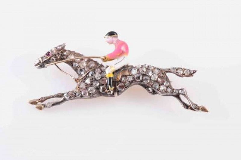 A DIAMOND SET ENAMEL HORSE AND JOCKEY BROOCH, CIRCA 1880, depicting the horse at full gallop, set throughout with graduating rose cut diamond highlights with a ruby set highlight for the eye, the jockey with black, white and pink enamel detailing. In fitt