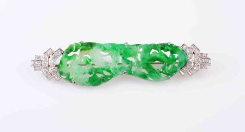 A JADEITE AND DIAMOND BROOCH, the heavily pierced and engraved jadeite plaque claw set in an open mount, set between structured diamond set end terminals, each set with sixteen brilliant cut slightly graduating diamonds, all set on a brooch pin fitting. L