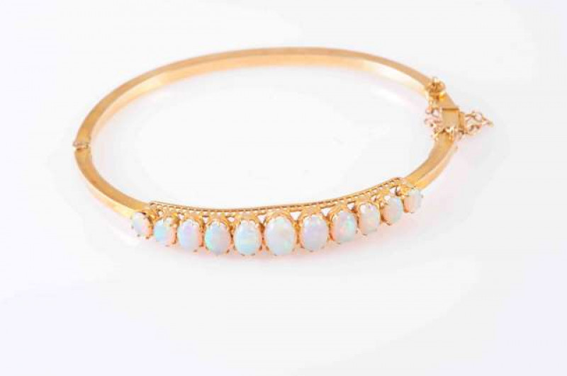 A VICTORIAN OPAL AND GOLD BANGLE, CIRCA 1890, the graduating row of eleven oval cabochon-cut opals claw set above pierced mount, on plain narrow bangle, with integral clasp, stamped '15ct'. With safety chain. Internal diameter 6cm. Total weight approximat