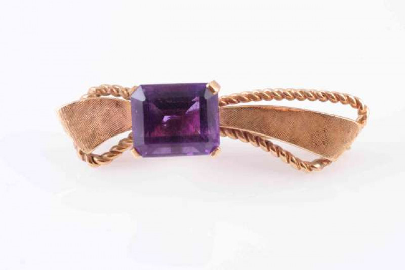 AN AMETHYST BROOCH, the square cut amethyst four claw set to the off-centre of a bow design mount of scroll work and brushed metal detailing, on brooch pin fitting. Stamped 10K. Makers mark GAJ. Length approximately 4.5cm. Weight 7.3gms.