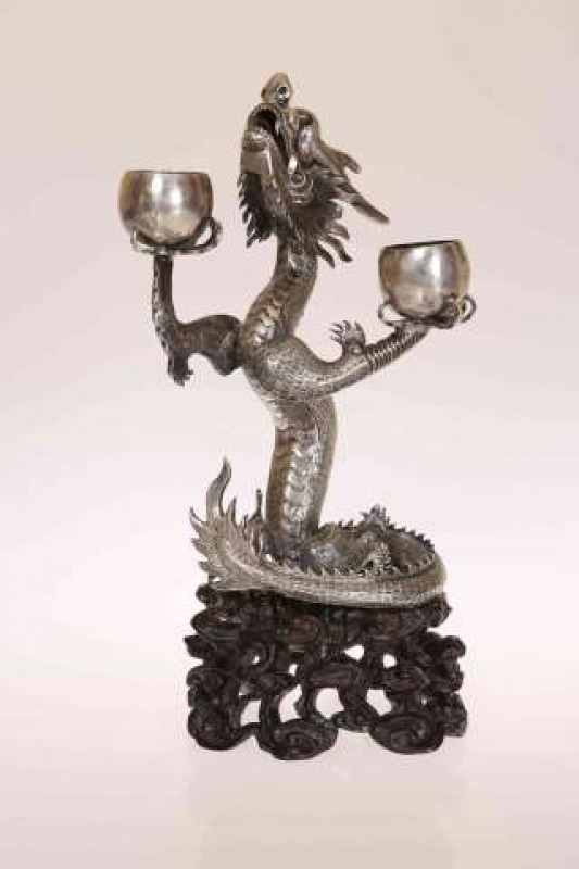 A CHINESE EXPORT SILVER CANDLESTICK, Hung Chong, in the form of a five-clawed dragon holding aloft two candlecups, it's sinuous body raised on a carved and pierced wood stand, struck HC twice. 22.5cm overall