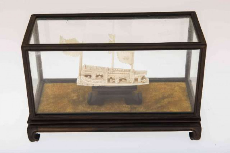 A CHINESE HARDWOOD AND GLASS DISPLAY CASE CONTAINING A MINIATURE IVORY BOAT, with figures, masts and flags, circa 1900. Case length 25cm