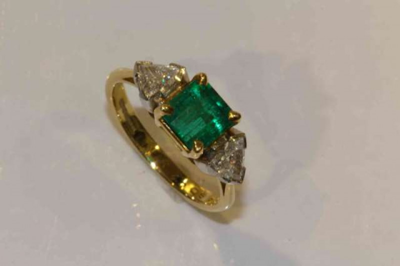 An emerald and diamond ring, the square cut emerald four claw set between two trillion cut claw set diamonds, all set in a simple open mount between open tapering shoulders on a plain band, stamped 750, ring size P, weight 5gms