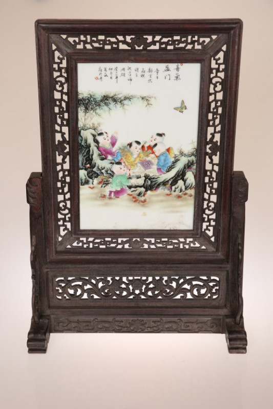 A CHINESE PORCELAIN TABLE SCREEN, painted with children at play in a landscape, within a fret-carved wooden frame and stand. 66cm by 45cm