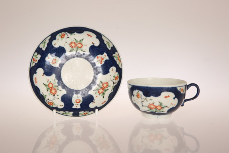 A WORCESTER SCALE BLUE GROUND CUP AND SAUCER, CIRCA 1770, enamelled in Kakiemon style with panels of chrysanthemum and other flowers, square seal mark. Saucer 13cm