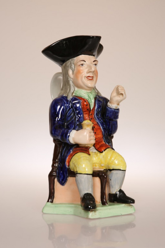 A STAFFORDSHIRE TOBY JUG OF THIN MAN TYPE, CIRCA 1840, seated on a chair with a tricorn hat and holding a jug, numbered 380 to base. 28cm