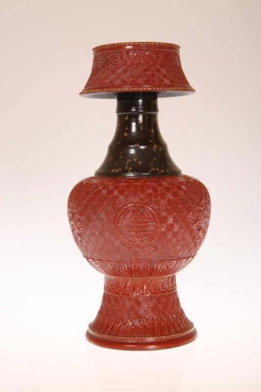 A CHINESE PORCELAIN CANDLE HOLDER, glazed and moulded to simulate cinnabar lacquer, of baluster form, with waisted candle cup above a black and gilt neck, and further decorated with Chinese symbols, gilt seal mark to base, in a fitted velvet-lined case. 1
