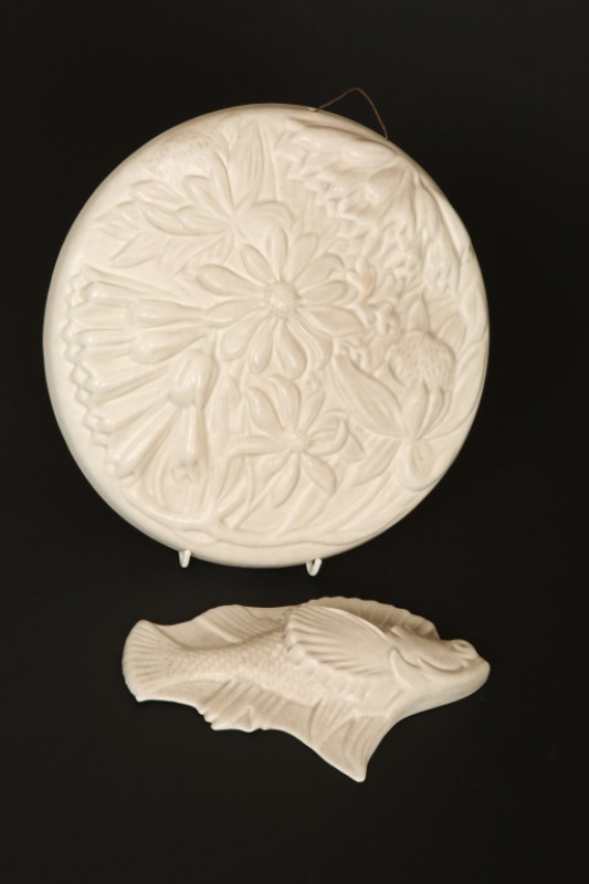 TWO PILKINGTONS ROYAL LANCASTRIAN WALL PLAQUES, second quarter 20th Century, each in an ivory glaze, the first modelled as a fish, the second circular modelled with flowers and foliage, each with impressed mark. 21cm and 27cm