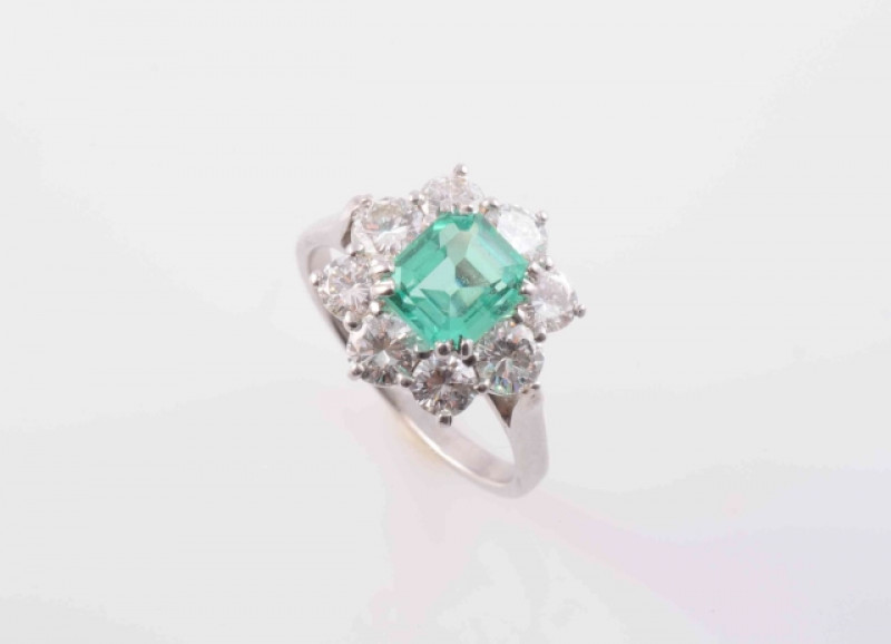A COLOMBIAN EMERALD AND DIAMOND RING, the square cut emerald four double claw set raised within a surround of eight brilliant cut diamonds in raised pierced mount between tapering pierced shoulders on a plain band. Ring size N. Weight 4.3 grams.  Accompan