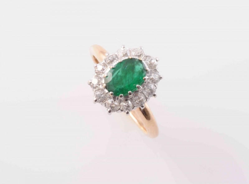 AN EMERALD AND DIAMOND RING, the oval cut emerald twelve claw set within a surround of twelve brilliant cut diamonds in an open pierced detail mount between solid tapering shoulders on a plain band. Ring size L. Weight 3.8 grams.