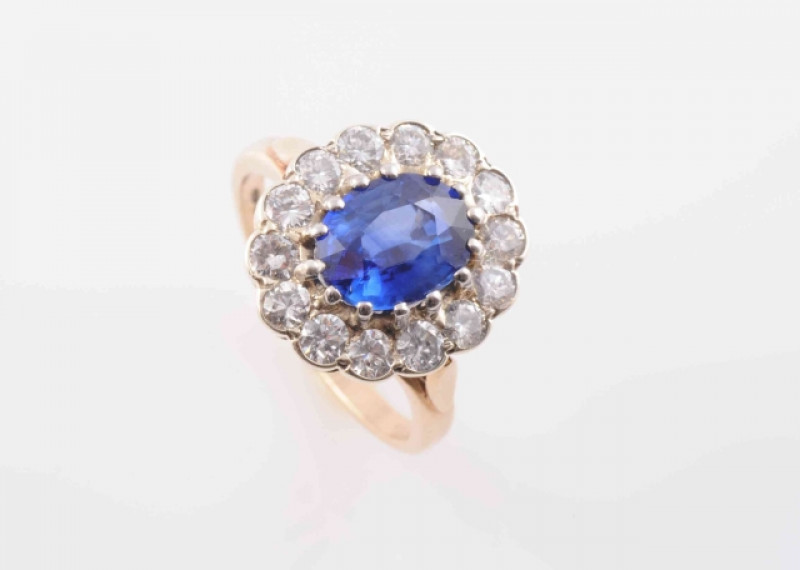 A SAPPHIRE AND DIAMOND RING, the oval cut sapphire fourteen claw set within a surround of fourteen brilliant cut diamonds in scalloped edge mount, between pierced tapering shoulders on a plain band. Stamped 375. Ring size M. Weight 4.2 grams.