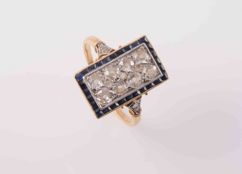 AN EDWARDIAN SAPPHIRE AND DIAMOND RING, the rectangular mount set with eleven graduating old brilliant cut diamonds within a surround of square cut sapphire highlights, all set in an open pierced detail mount between pierced and moulded detail shoulders e